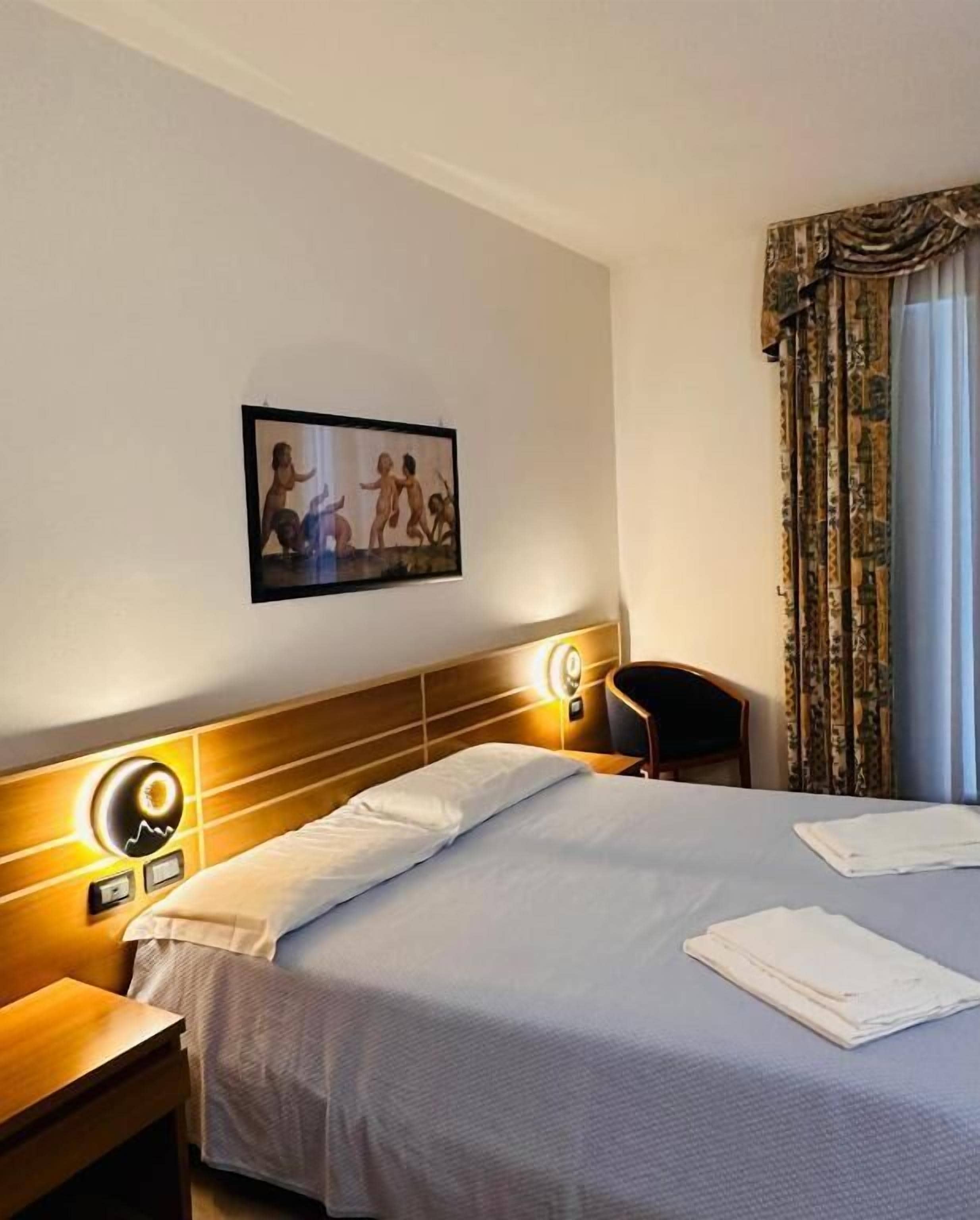 Habitación de hotel con cama, mesita de noche, silla, cuadro en la pared y ventana con cortinas.