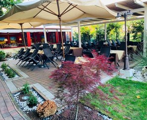 Un área de comedor al aire libre con mesas y sillas, rodeada de un jardín con diversas plantas y un sendero.