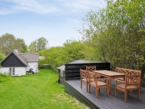 Una terraza de madera con una mesa y sillas, un cobertizo pequeño y un edificio blanco de techo oscuro.