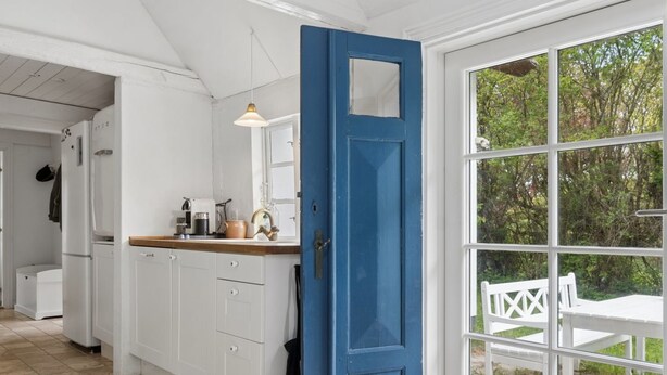 Een keuken met witte kasten, een blauwe deur en een raam met uitzicht op groen.