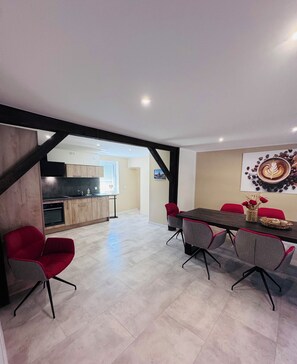 A modern interior with a dining area featuring a dark wooden table and red chairs, a kitchen with wooden cabinets, and a large window. - Elementa Lodges Wernigerode-Reddeber (Reddeber)