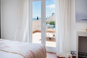 Une chambre à coucher avec un lit, des rideaux blancs, une vue sur la mer et un balcon avec une petite table.