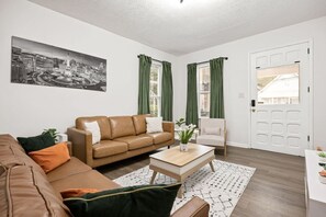 Ein Wohnzimmer mit braunem Ledersofa, einem weißen Couchtisch, einem Teppich mit grün-weißen Mustern und einem Stadtbild an der Wand.