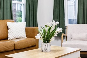 Una sala de estar con un sofá marrón, un sillón blanco, una mesa de centro de madera y un florero con flores blancas.