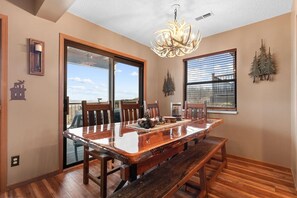 A dining area with a wooden table, benches, a chandelier, and a view through large windows.