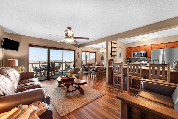A spacious living room with a large sofa, a dining area with wooden chairs, a kitchen with stainless steel appliances, and a balcony with a view.