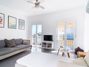 Ein modernes Wohnzimmer mit einem grauen Sofa, einem weißen Couchtisch und einem Flachbildfernseher auf einem weißen Schrank. Zwei Türen führen zu einem Balkon mit Meerblick.