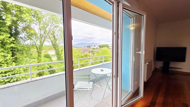 Een balkon met een tafel en stoelen, een televisie en uitzicht op groen en gebouwen.