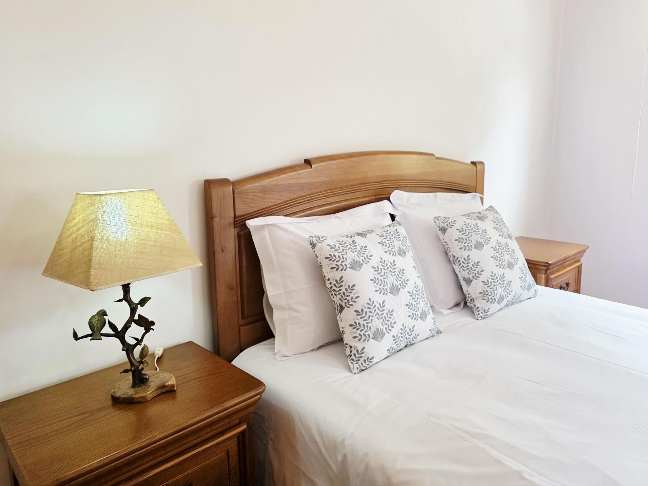 Un lit bien fait, avec une literie blanche et des coussins à motifs, une table de chevet en bois avec une lampe, et un petit arbre décoratif.