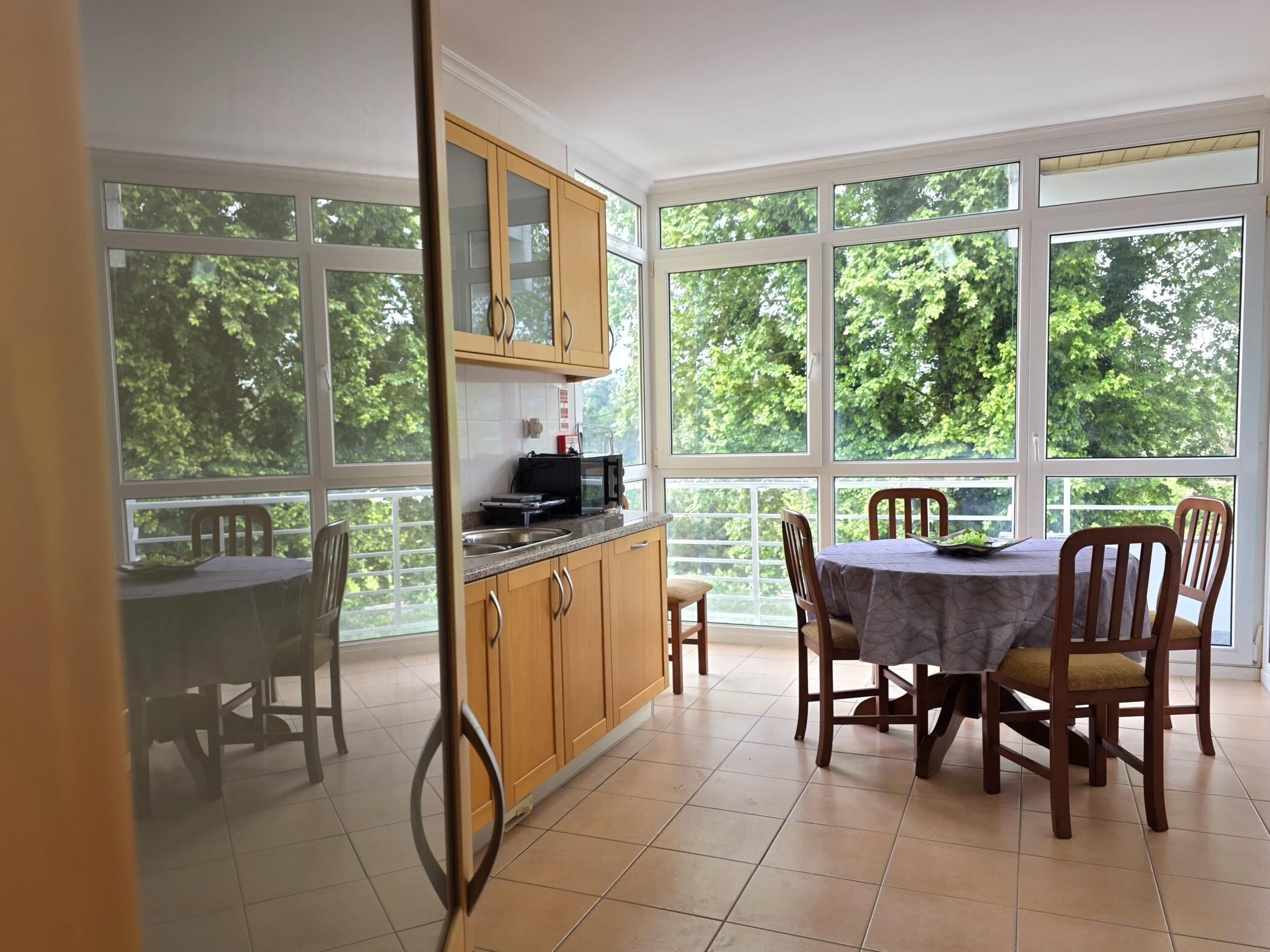 Een eetgedeelte met een tafel en stoelen, een keuken met kasten en een magnetron, en grote ramen met uitzicht op bomen.