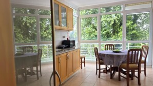 Een eetgedeelte met een tafel en stoelen, een keuken met kasten en een magnetron, en grote ramen met uitzicht op bomen.