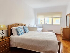 A bedroom with a wooden bed, white bedding, a bedside lamp, a dresser, and a window with blinds. - Andorinhas Apartment | Monção | Wine Tourism | Town Centre (Monção)
