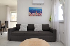 A modern living room with a grey sofa, a painting of a coastal scene, and a dining area in the background. - Nice 2 Bed Apartment (Okrug)