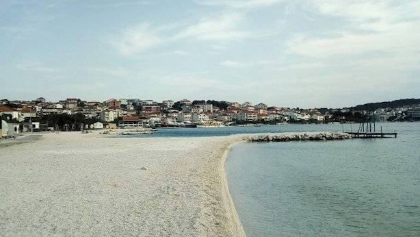Ein Küstenstrand mit freiem Blick auf die Uferlinie, einer in das Wasser ragenden Mole und einer Stadt im Hintergrund.