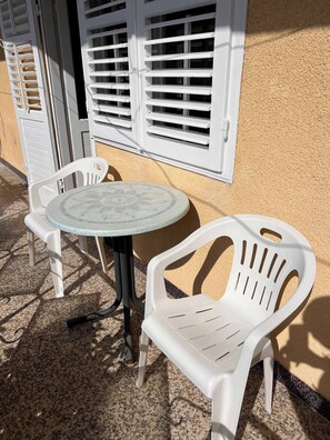 A small outdoor seating area with a round table and two chairs, a window with shutters, and a textured wall. - Sunny Apartment in Trogir (Trogir)
