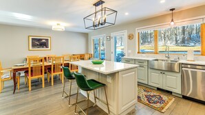 Una cocina moderna con una isla central, taburetes de bar, un comedor con sillas de madera, un amplio ventanal con vistas y un lavamanos de acero inoxidable.