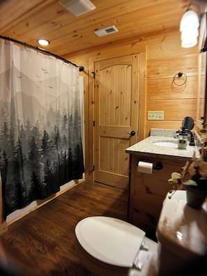 A wooden cabin bathroom with a shower curtain depicting a forest scene, a toilet, a sink with a mirror, and a small vase with flowers.