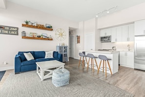 Una sala de estar moderna con un sofá azul, una mesa de centro blanca y una cocina con alacenas blancas y un refrigerador de acero inoxidable.