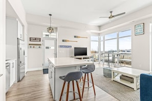Una cocina moderna con una isla central, taburetes de bar, un comedor con mesa y sillas, y ventanas amplias que ofrecen vistas al exterior.