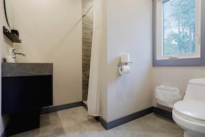 A bathroom with a toilet, a shower curtain, a towel dispenser, and a window with a view of trees. - 3-Bedroom Cottage #10 (Nobel)