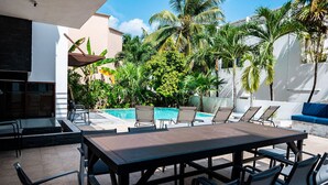 Ein Poolbereich mit Esstisch und Stühlen, eine Lounge-Ecke mit einer Couch und ein Pool, der von Palmen umgeben ist.