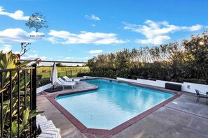 A swimming pool with lounge chairs, a small table, and a view of the countryside.