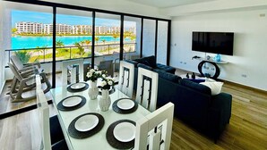 Ein modernes Wohnzimmer mit Essbereich, großen Fenstern, einem Flachbildfernseher und Blick auf ein Resort und den Ozean.