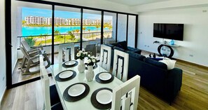A modern living room with a dining area, large windows, a flat-screen TV, and a view of a resort and ocean. - Urban Oasis- Modern comfort and stunning beach views (Punta Cana)
