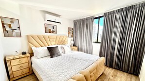 Ein Schlafzimmer mit einem gesteppten Kopfteil, einem Bett mit einer gesteckten Bettdecke, einem Nachttisch mit Schubladen, einer Klimaanlage und einem Fenster mit Vorhängen.