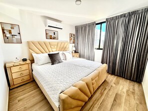 A bedroom with a tufted headboard, a bed with a quilted cover, a nightstand with drawers, an air conditioning unit, and a window with curtains. - Urban Oasis- Modern comfort and stunning beach views (Punta Cana)