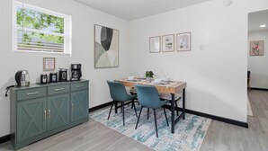 Een eetgedeelte met een tafel voor vier personen, twee stoelen, een kast met lades, een raam met zonwering en lijstwerken aan de muur.