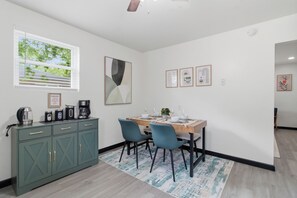 Uma sala de jantar com uma mesa posta para quatro pessoas, duas cadeiras, um armário com gavetas, uma janela com persianas e obras de arte emolduradas na parede.