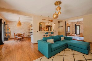 A spacious living area with a teal sectional sofa, wooden dining table, and a kitchen visible in the background.