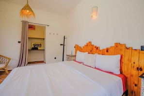 A bedroom with a wooden headboard, a bed with white bedding, a wicker chair, and a door leading to a bathroom.