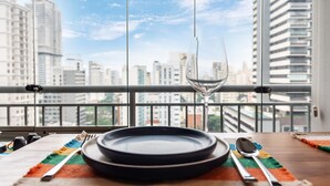 Une table à manger avec une assiette, un verre et des couverts, le tout posé sur un set de table coloré, avec en arrière-plan une vue sur la ville, visible à travers la rambarde d’un balcon.
