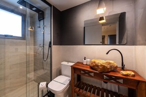 Un baño moderno con cabina de ducha de vidrio, un inodoro blanco, un tocador de madera con una gran roca y un espejo sobre el lavabo.