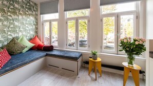 Een kamer met een ingebouwde bank, grote ramen met zonwering, een klein rond tafeltje met een plant en een vaas met bloemen.