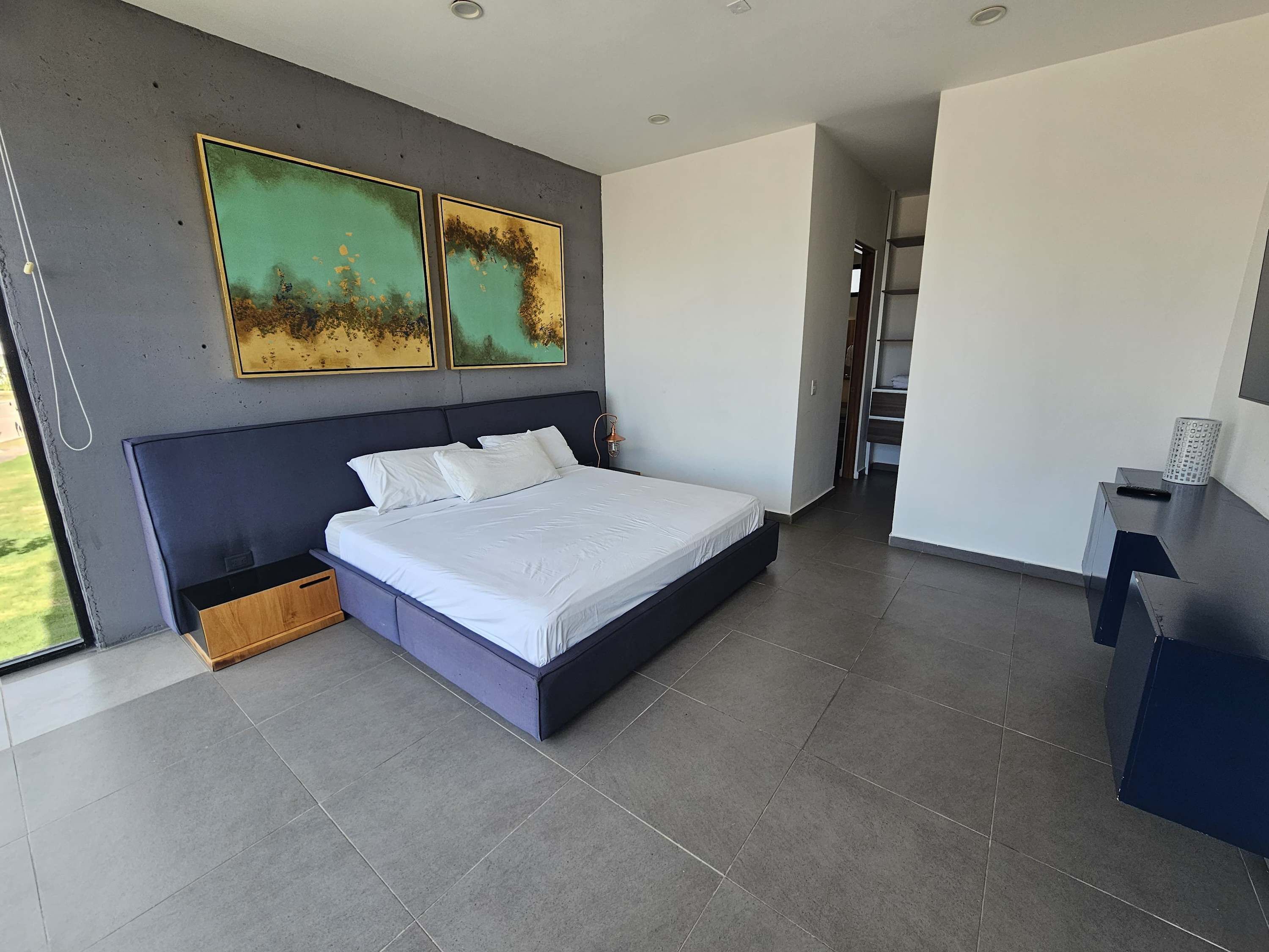 Une chambre à coucher moderne avec un grand lit, une table de chevet, un bureau et deux tableaux abstraits accrochés au mur.