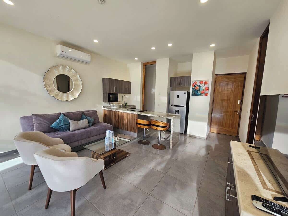 A modern living room with a sofa, two armchairs, a coffee table, and a kitchen area with a refrigerator, microwave, and bar stools.