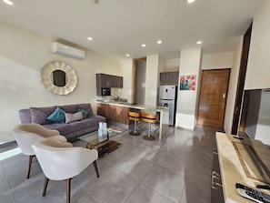 Una sala de estar moderna con un sofá, dos sillones, una mesa de centro y una cocina con refrigerador, microondas y taburetes de bar.