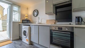 A compact kitchen with a washing machine, oven, microwave, and a clock on the wall. - Tide & Seek — Garden Flat by the Sea (Walmer)