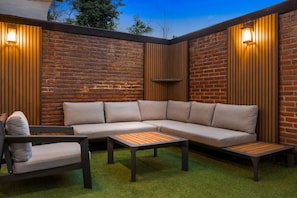A sectional sofa, two wooden tables, and a chair in an outdoor patio area with brick walls and vertical wooden slats.