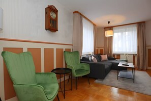Ein Wohnzimmer mit einer Couch, zwei grünen Sesseln, einem kleinen Tisch, einer Wanduhr und einem Fenster mit Vorhängen.