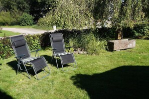 Zwei Gartenliegen auf einer Rasenfläche, mit einem Steinpflanzgefäß und dichtem Grün im Hintergrund.