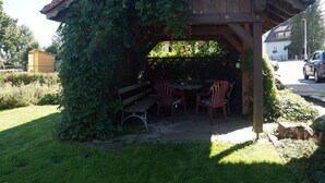 Ein hölzerner Pavillon mit Tisch und Stühlen, umgeben von viel Grün.