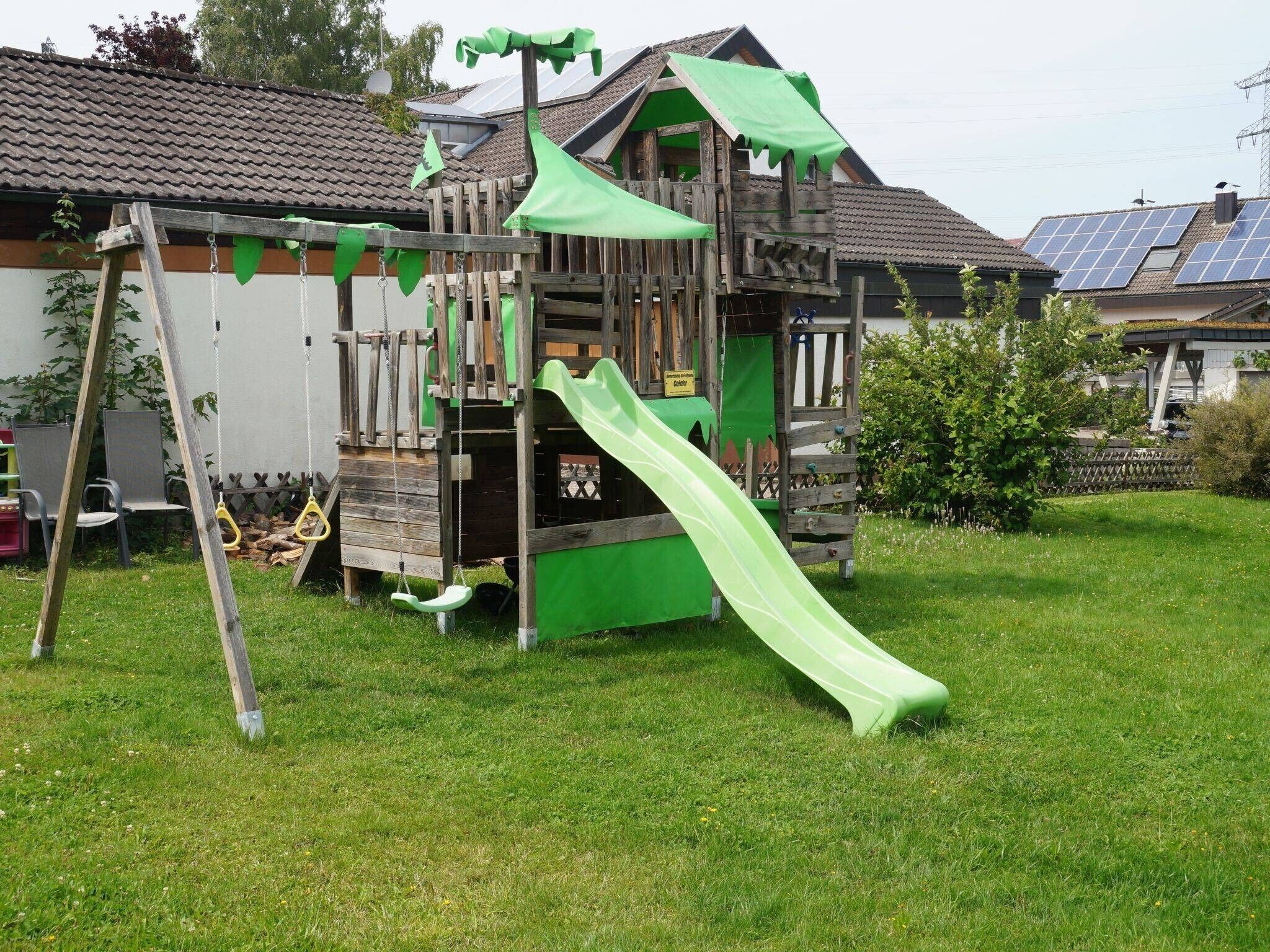 A wooden playhouse with a slide, a swing set, and a climbing wall.