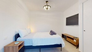 Ein Schlafzimmer mit einem Bett, einem Fernseher, einem Holznachttisch und einer Deckenleuchte.