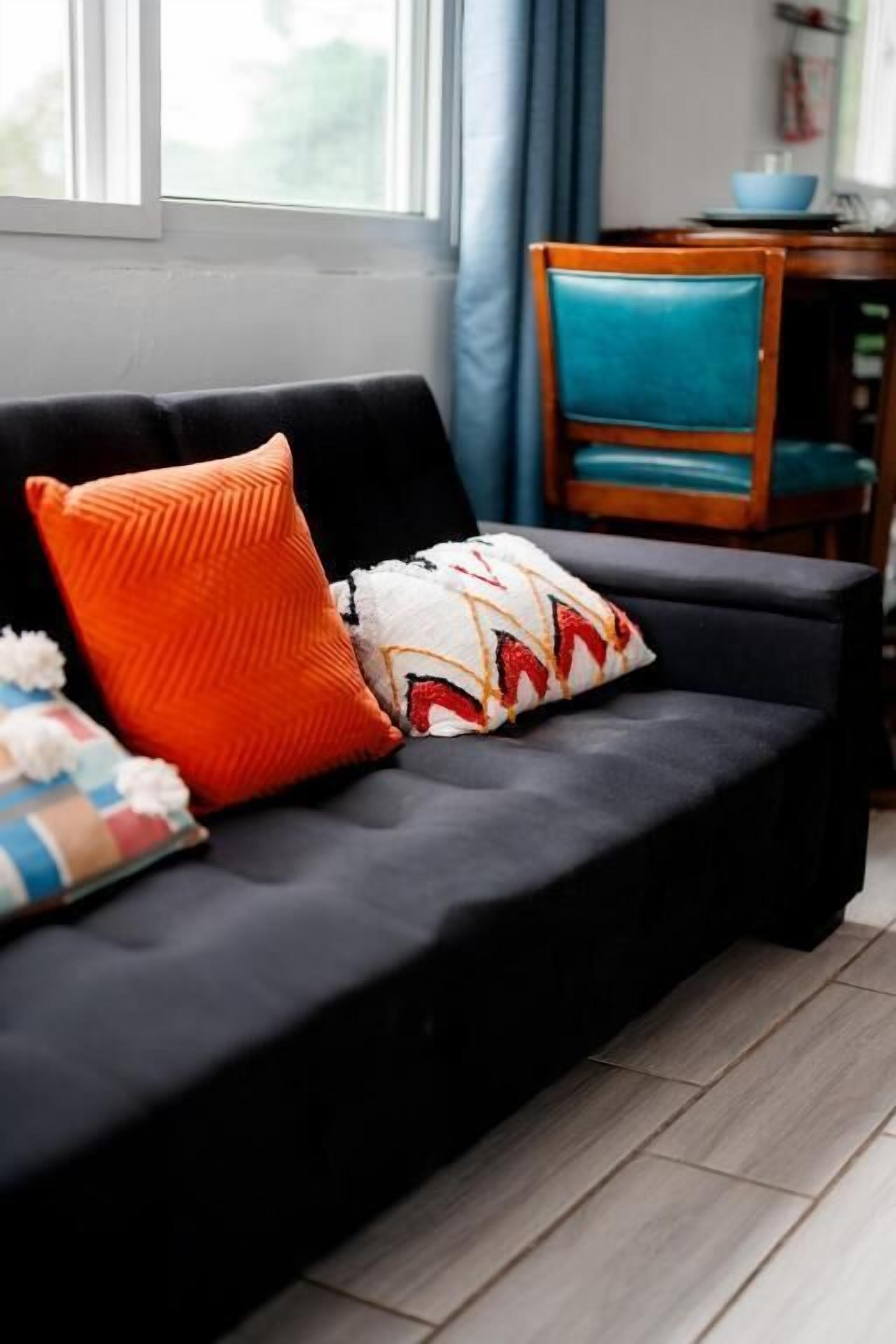 A dark-colored sofa with two pillows, a wooden chair, and a dining table with a blue bowl.