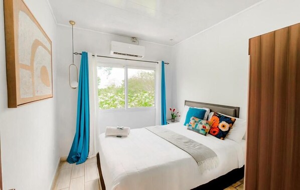 Un dormitorio con cama, ventana, cortinas, un equipo de aire acondicionado y un cuadro en la pared.