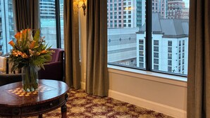 A hotel room with a large window, a chandelier, a vase with flowers, and a patterned carpet.